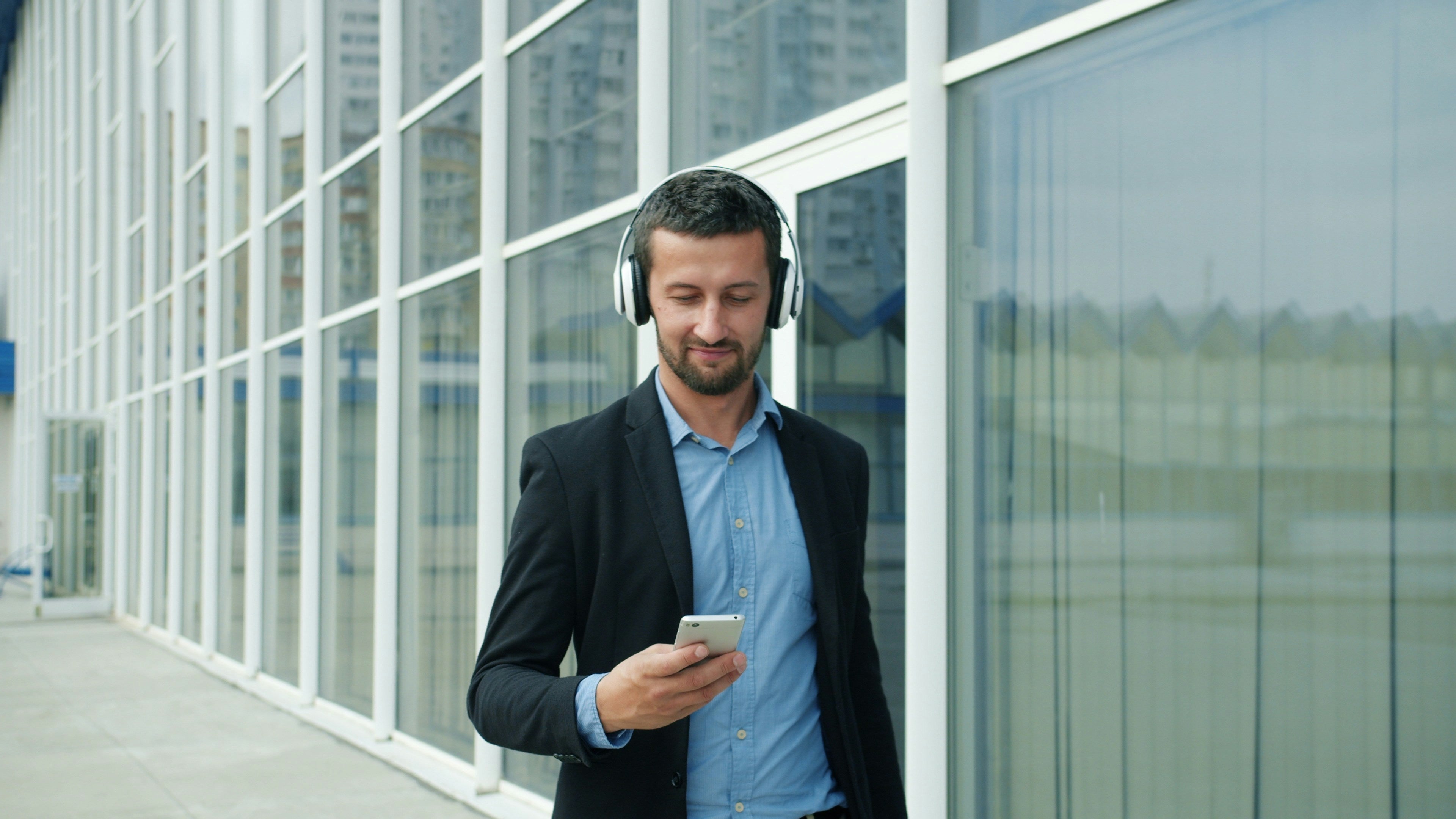 A man in a suit and blue shirt takes a walk outside in front of his office and reads his phone with headphones on